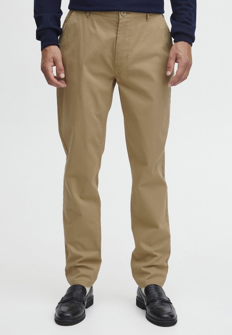 Chinos khaki avec une coupe droite, fabriqués en tissu lisse, dotés d'une fermeture à bouton et de poches avant. Portés avec des mocassins noirs.
