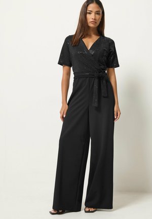 Femme portant une combinaison noire à paillettes avec un haut portefeuille, manches courtes et pantalons larges, debout devant un fond clair uni.