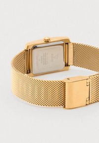 Rechthoekig gouden horloge met een mesh band, metalen afwerking, een doorzichtige glazen achterkant en een gestructureerde band met een sluiting.