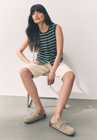 Haut sans manches à rayures vertes, short beige clair et sabots en daim gris avec semelle en liège. Assis sur une chaise en métal devant un fond neutre.