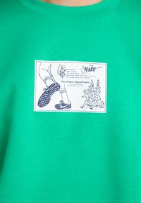 Felpa verde in cotone con un patch grafico che raffigura corridori, alberi e il logo Nike. Design classico con scollatura rotonda e colletto a costine.
