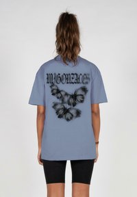 Lyseblå bomulds t-shirt med et grafisk design af sorte sommerfugle og teksten "MIGONZALES" på ryggen. Afslappet pasform, korte ærmer.