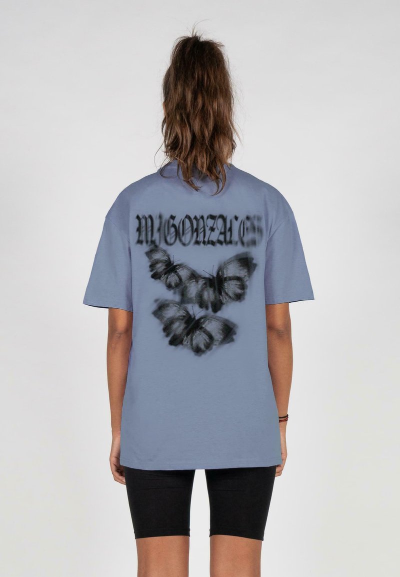 Lyseblå bomulds t-shirt med et grafisk design af sorte sommerfugle og teksten "MIGONZALES" på ryggen. Afslappet pasform, korte ærmer.
