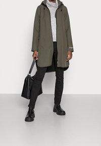 Olivengrøn lang jakke med hætte, lynlås og sidelommer. Båret over en grå striktrøje, kombineret med sorte bukser og sorte støvler.