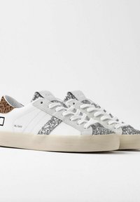 Hvide sneakers med sølvglitterdetaljer, struktureret dyreprint hæl, flade hvide snørebånd og en beige gummisål. Simpelt design med branding.