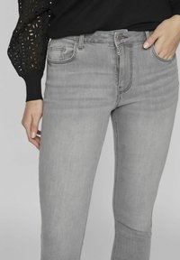 Los vaqueros de mezclilla gris presentan un diseño ajustado con cremallera y bolsillos frontales, combinados con una blusa negra texturizada.