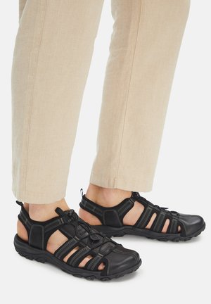 Schwarze Sandalen aus Synthetik mit offener Zehenpartie, verstellbaren Riemen und strukturierten Sohlen, kombiniert mit hellbeigen Hosen.