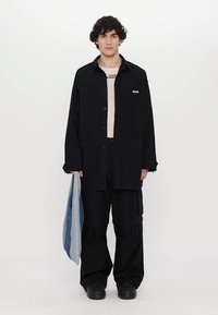 Camisa negra de gran tamaño y pantalones cargo hechos de una tela con acabado mate, sobre una camisa de color claro, sosteniendo una chaqueta de mezclilla azul.