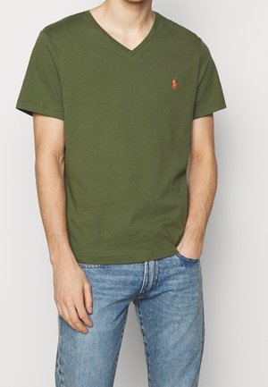 Mann trägt olivgrünes V-Ausschnitt-T-Shirt mit kleinem orangefarbenem Logo auf der linken Brust und hellblaue Jeans, Hände entspannt, eine in der Tasche.