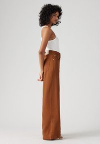 Korkeavyötäröiset ruskeat laidasta levenevät housut, jotka on valmistettu tukevasta kankaasta ja joissa on suora leikkaus, takataskut sekä hopeansävyiset yksityiskohdat.