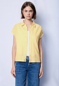 Camicia gialla a maniche corte con bottoni, realizzata in un tessuto leggero, con colletto e vestibilità rilassata. Indossata sopra un top bianco a spalla.