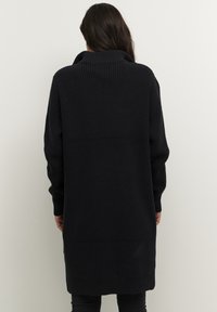 Cardigan noir long en tricot avec un col haut, texture côtelée et manches longues. Présente une coupe droite et tombe sous le genou.