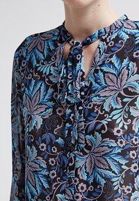 Svart blommig blus med blå och lavendelbladsmönster, vertikal knytning vid halsringningen och knappstängning framtill.
