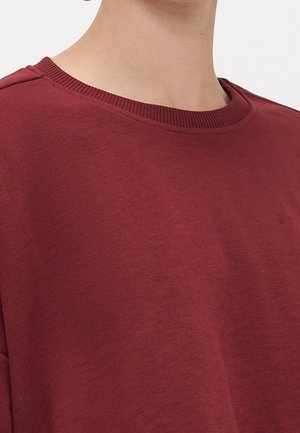 Gros plan d'une personne portant un sweat-shirt uni rouge foncé à col rond, montrant le cou et la partie supérieure de la poitrine.