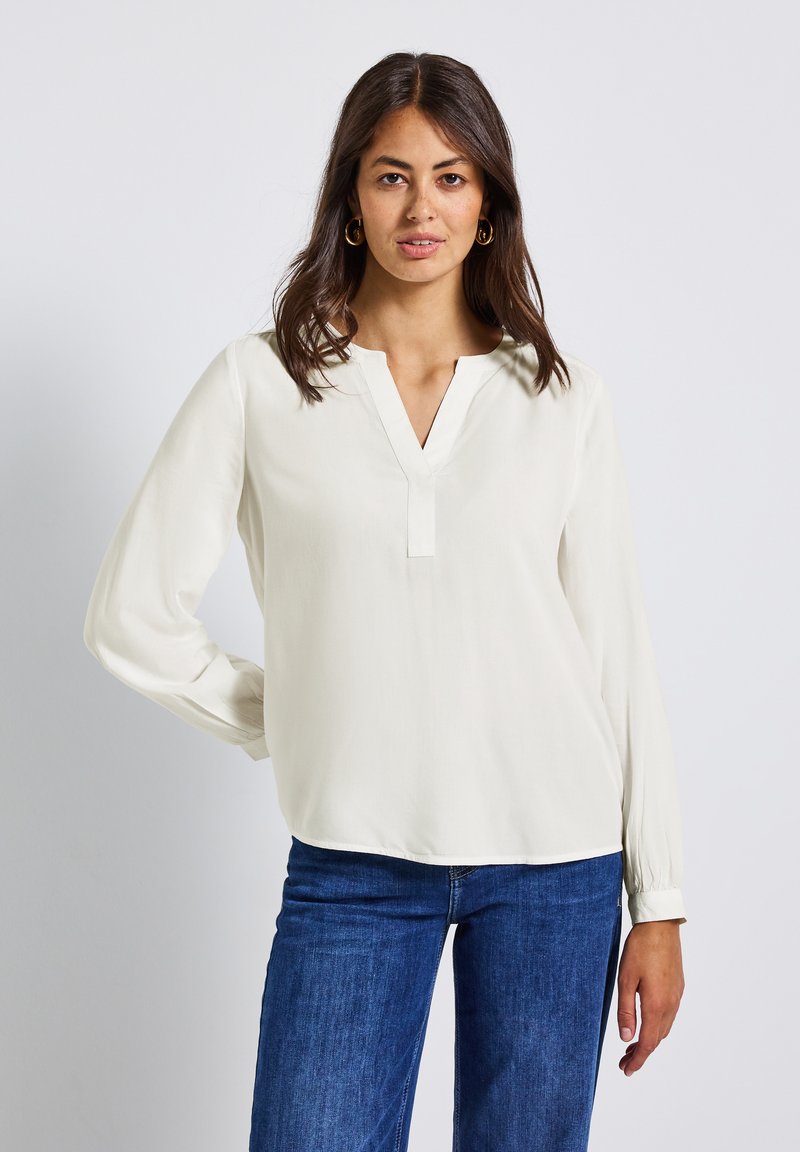 Femme aux longs cheveux noirs, portant de grandes boucles d'oreilles en or, une blouse blanche à manches longues et décolleté en V, ainsi que des jeans bleus, se tenant devant un fond uni.