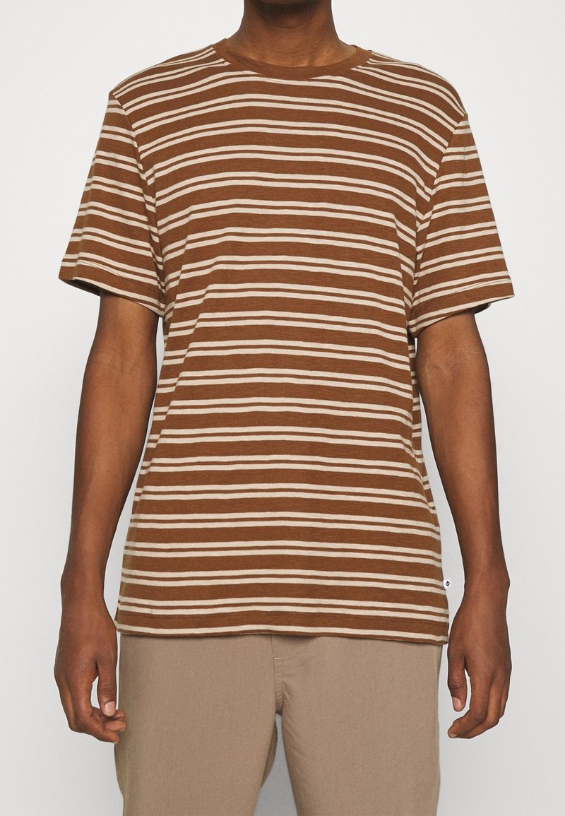 Homme portant un t-shirt à manches courtes rayé horizontalement marron et beige avec un pantalon beige, debout devant un fond uni.