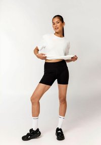 Junge Frau in weißem T-Shirt, schwarzen Radhosen, weißen Socken und schwarzen Sneakers zieht ihr Shirt leicht hoch vor einem schlichten Hintergrund.