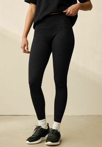 Sorte ribbede leggings lavet af strækbart materiale, tætsiddende design, høj talje, parret med sorte sneakers og hvide ankelstrømper.