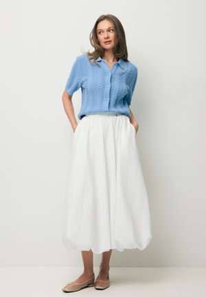 Vrouw in een blauw shirt met korte mouwen en kabelgebreide structuur, witte broek met wijde pijpen en beige platte schoenen, staand met handen in de zakken tegen een effen achtergrond.