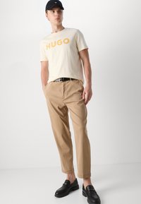 T-shirt en coton beige avec "HUGO" en orange, associé à un pantalon marron clair. Mocassins noirs avec des accents en matériaux dorés. Casquette noire.