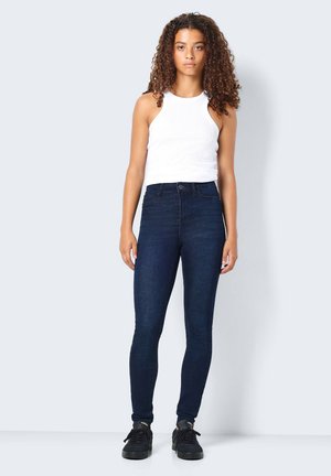 Jeans skinny de mezclilla azul oscuro con cintura alta, que presentan un diseño clásico de cinco bolsillos y un desvanecimiento sutil. Llevados con una blusa sin mangas blanca.