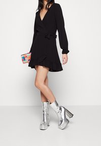 Robe noire à manches longues et ourlet volanté, associée à des bottes plateforme métalliques argentées et à une pochette à motifs multicolores.