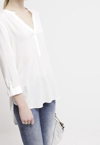 Hvid, langærmet bluse med V-hals og knapdetaljer, lavet af letvægtsstof. Båret sammen med blå jeans og en hvid taske.