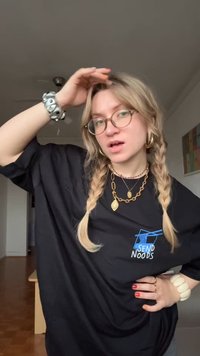 Overdimensioneret sort T-shirt med "SEND NOODS" grafik, båret med lagdelte guldkæder, forskellige armbånd og flettet hår.