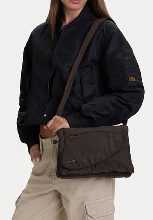 Mujer con una chaqueta bomber negra y pantalones cargo beige, llevando un bolso de hombro G-Star marrón oscuro cruzado sobre su cuerpo.