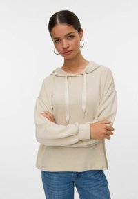 Beige mikina s měkkou texturou, kapucí na šňůrku a přehozenými dlouhými rukávy. V kombinaci s modrými džínami model pózuje se zkříženýma rukama.