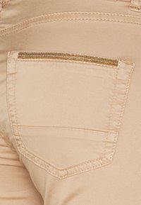 Beige fabric pocket on pants with a thin gold beaded accent at the top. Features contrasting stitching and a classic shape.