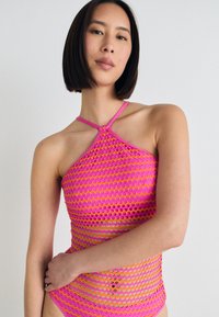 Růžový síťovaný halter top s diamantovým vzorem, který má kontrastní design ramínek a zakulacený výstřih. Textura vypadá lehce a prodyšně.