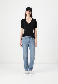 Sort kortærmet top med V-udskæring, lyseblå let tilpassede jeans og hvide sneakers. Modellen bærer en sort kasket.