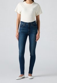 Camiseta ajustada de color beige claro y jeans ajustados de un azul oscuro. Los jeans tienen un diseño clásico de cinco bolsillos y un ligero desgaste. Zapatillas blancas sin cordones.