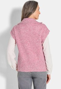 Cardigan en tricot rose avec une texture côtelée et un large col ; manches courtes sur une chemise à manches longues blanche ; associé à un jean gris.