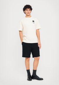 Beige t-shirt with short sleeves and black graphic design at the chest, paired with black shorts, dark socks, and black shoes.