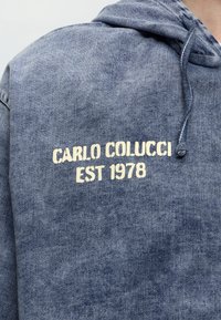 Μπλε φούτερ με υφή denim και λευκό κείμενο "CARLO COLUCCI EST 1978" στο πάνω μέρος της πλάτης, που φοράει ένα άτομο.