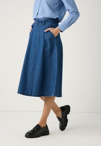 Frau trägt ein blaues, knöpfbares Hemd, einen hoch taillierten, mit Gürtel versehenen Jeans-Midi-Rock und schwarze, klobige Loafers, steht mit den Händen in den Taschen.