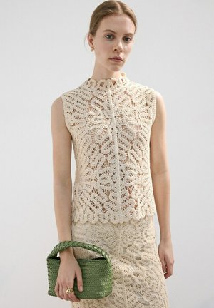 Mujer con top de crochet beige sin mangas y falda sosteniendo un pequeño bolso verde tejido contra un fondo gris liso.