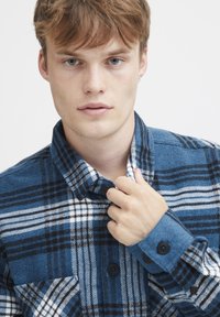 Blauwe en zwarte geruite flanellen shirt met witte accenten. Heeft een knoopsluiting bij de kraag en manchetten, met een zichtbaar borstzakje. Zachte textuur.