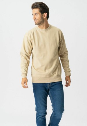 Mand med mørkt hår iført beige sweatshirt og blå jeans, stående og kiggende mod venstre foran en ensfarvet hvid baggrund.
