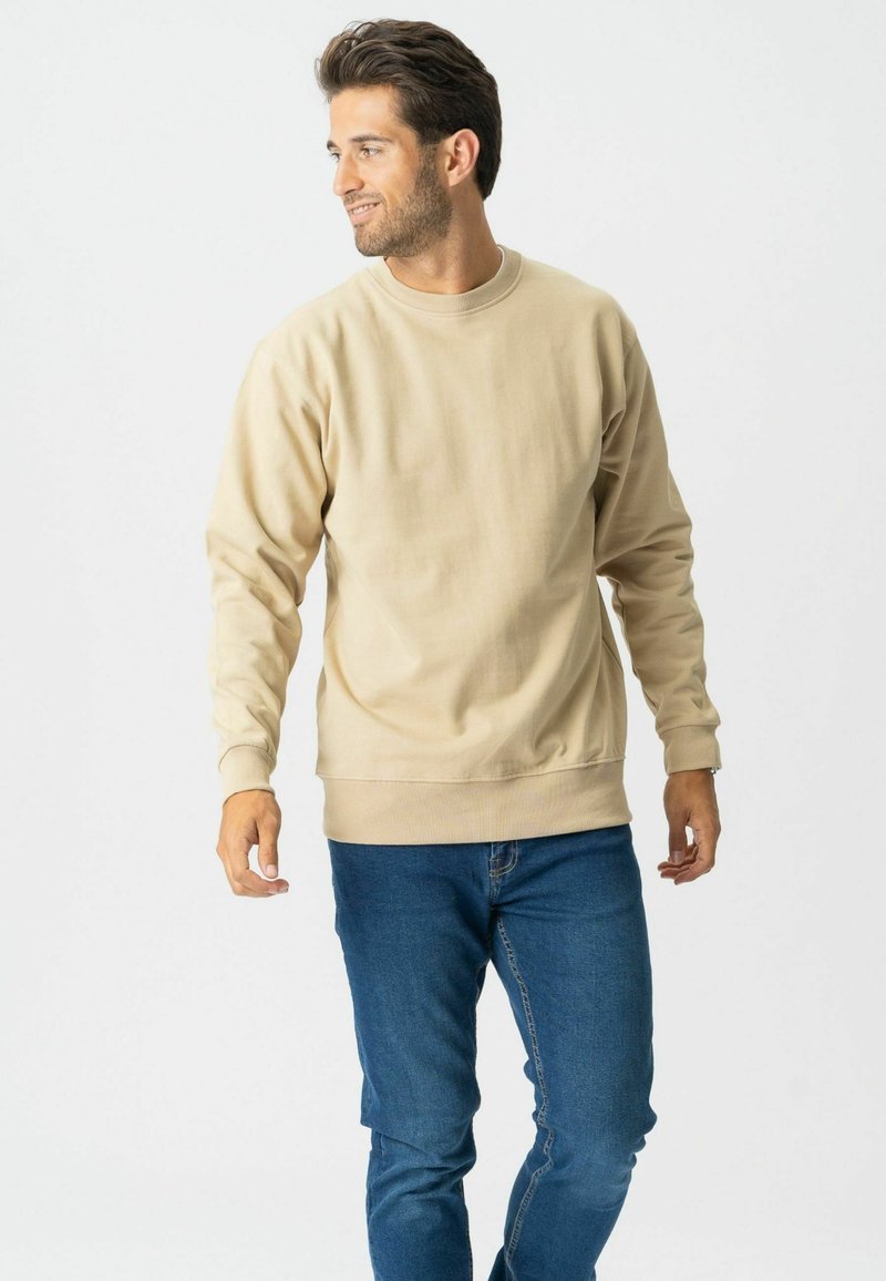 Homme aux cheveux foncés portant un sweat-shirt beige et un jean bleu, debout et regardant vers sa gauche sur un fond blanc uni.