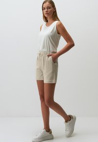 Witte mouwloze top en beige short met koordje. De outfit heeft een relaxte pasvorm en een textuurstof. Witte sneakers met gouden accenten.