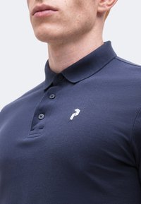 Hombre con una camiseta polo azul marino con un pequeño logo blanco con forma de flecha en el pecho izquierdo, recortado por debajo de la nariz.