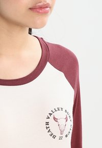 Raglanärmad skjorta med krämfärgad kropp och maroonärmar. Har en grafisk tryck av ett tjurhuvud och text på bröstet. Mjuk tygtextur.