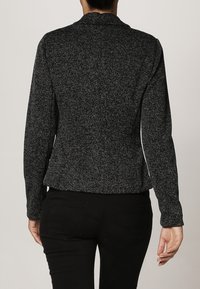 Blazer texturé gris foncé avec une silhouette ajustée, sans design particulier, et des manches longues. Subtiles accents blancs le long des coutures latérales.