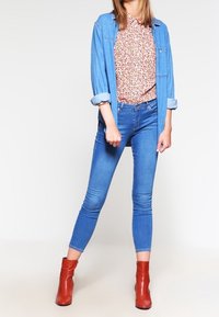 Donna che indossa una giacca di denim blu, una camicetta floreale, jeans skinny blu e stivali alla caviglia rossi, in piedi con le gambe divaricate su uno sfondo bianco.