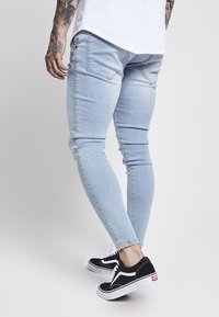 Vaqueros ajustados de color azul claro, elaborados en denim, con una textura suave y bolsillos traseros. Combinados con zapatillas negras con detalles en blanco.