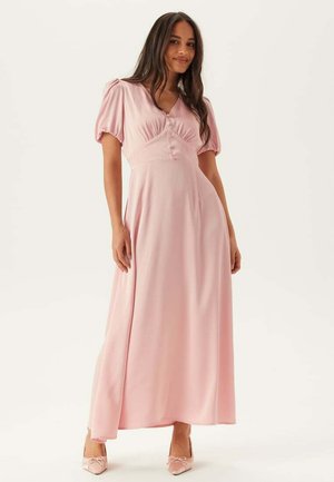 Femme portant une robe maxi rose clair à manches courtes bouffantes, boutonnée sur le devant, avec des talons pointus assortis, debout devant un fond neutre.