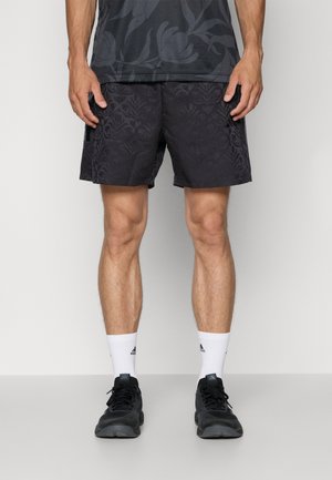 Homme portant un short noir à motif floral, des baskets noires et des chaussettes blanches Adidas, debout devant un fond gris clair.